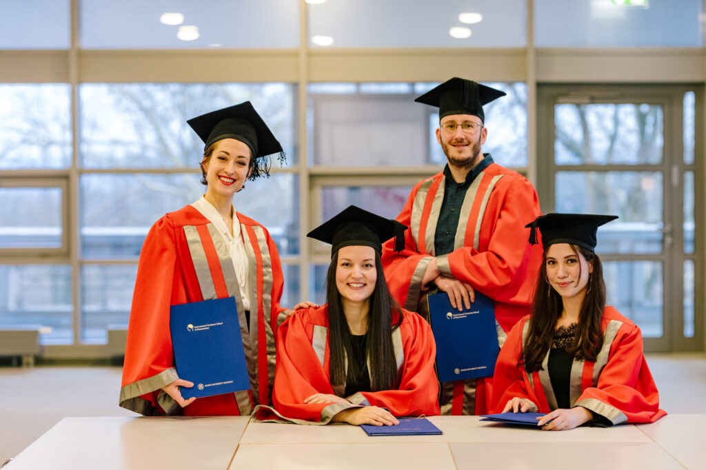 Four graduates in red and silver robes with black mortarboards pose with blue diploma folders. Two sit at a table while two stand behind them, smiling.