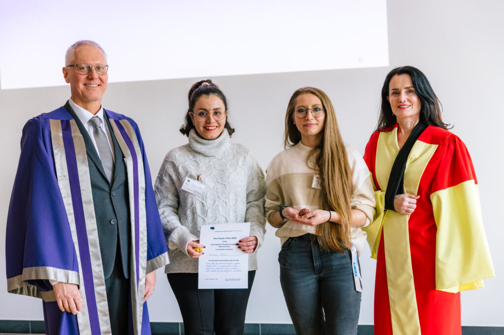 Four people pose for a photo at a graduation event, with one holding a certificate while two others wear academic robes.