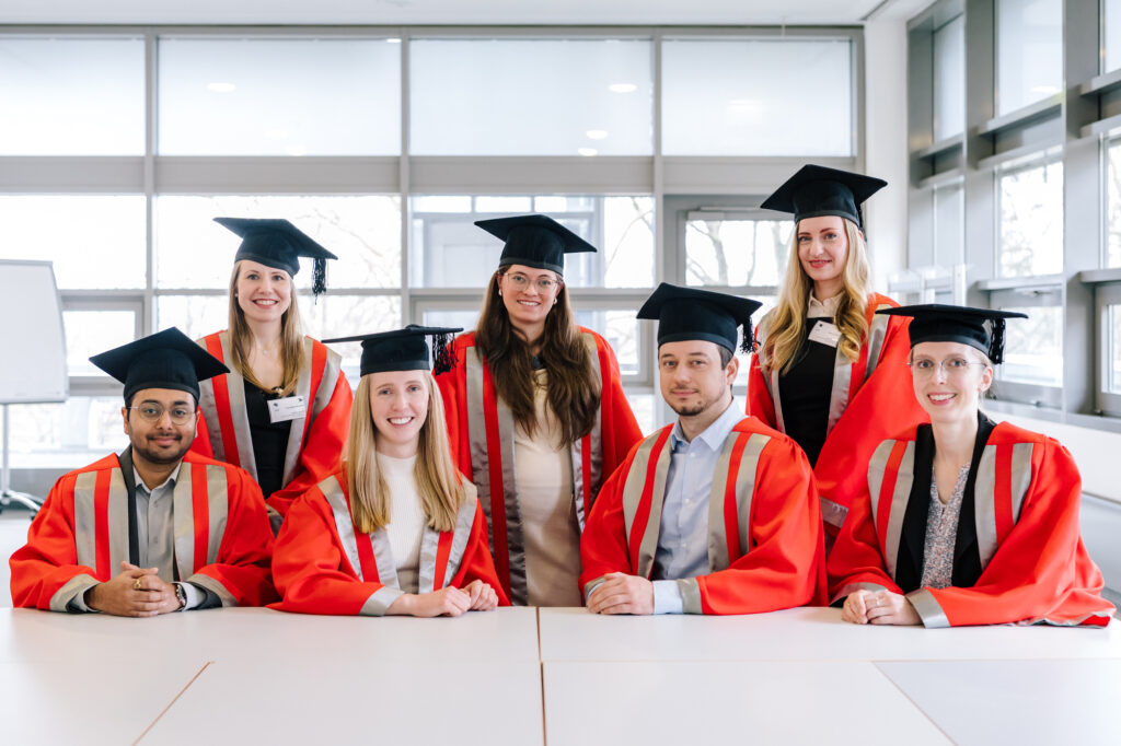 Eight graduates in red academic gowns and mortarboards pose together indoors during a graduation ceremony.