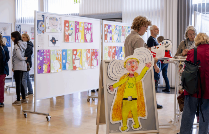 A previous Brain Day event showing an exhibition of children’s artwork. Visitors view colorful drawings displayed on panels, with a large cartoon figure labeled “Blitz Ohr” featured in the foreground.
