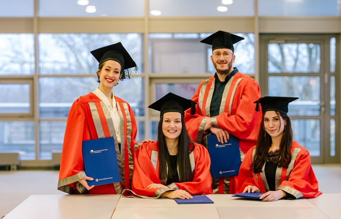 Four graduates in red and silver academic robes and black mortarboards pose with blue diploma folders. Two are seated at a table, while two stand behind them, smiling. The background features large windows with natural light.