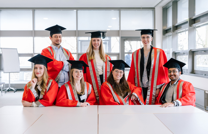 Eight graduates in red academic gowns and mortarboards pose together indoors during a graduation ceremony.