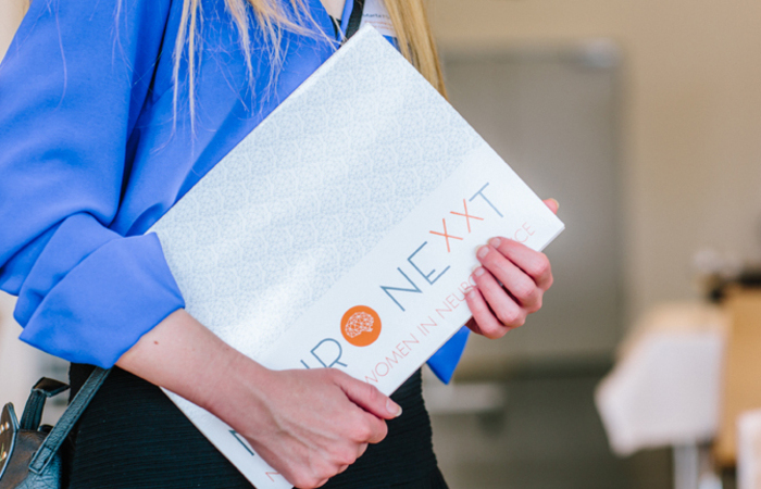 A person in a blue blouse holding a folder labeled 'NeuroNexxt: Women in Neuroscience.'
