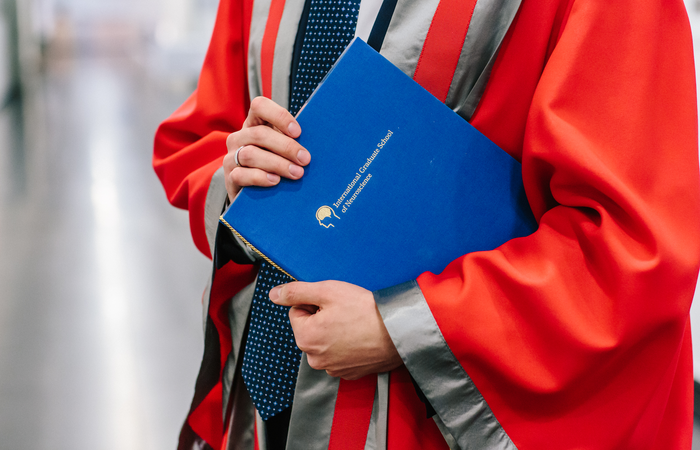 A person in a red and gray academic robe holding a blue folder labeled 'International Graduate School of Neuroscience.'