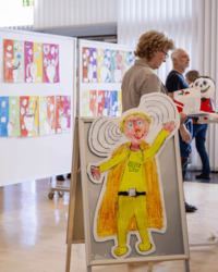 A past Brain Day event showing visitors viewing colorful children’s drawings on display panels. In the foreground stands a large illustrated cutout of a superhero-like figure labeled “Blitz Ohr.”