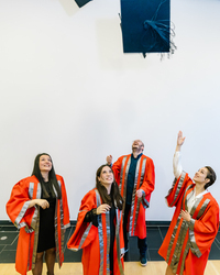 Four graduates in red and silver academic robes celebrate by throwing a black mortarboard into the air. They are smiling and looking up, marking the end of their PhD journey.